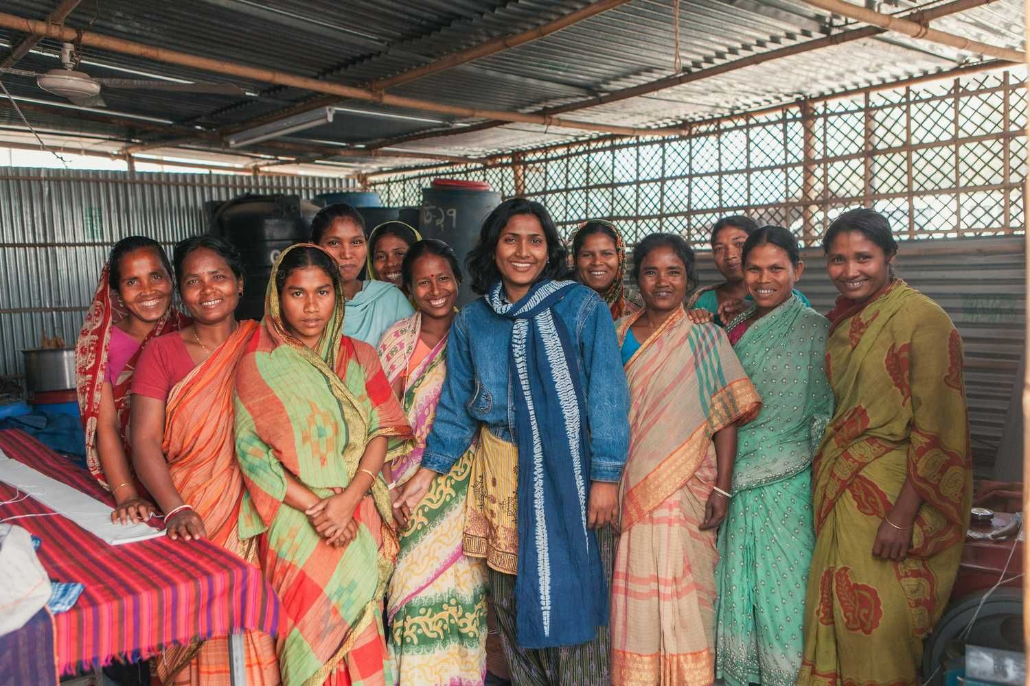 A group of smiling women, most wearing colorful saris, stand together inside a simple corrugated metal structure.