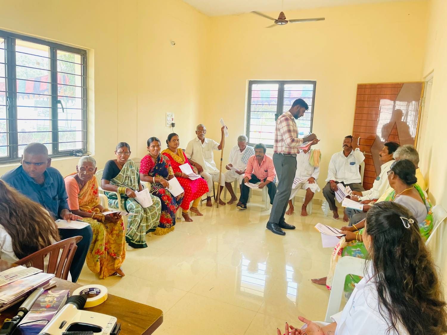 Community health awareness session with participants gathered indoors, receiving guidance and information from a facilitator