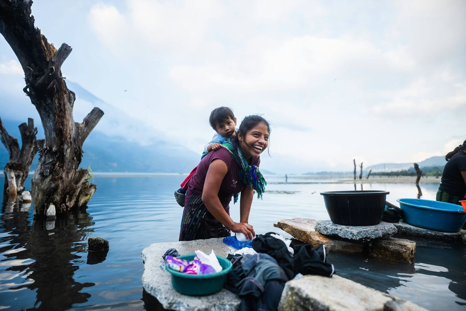 A smiling woman washes laundry by a lake with a young child perched on her back against a backdrop of mountains and misty water