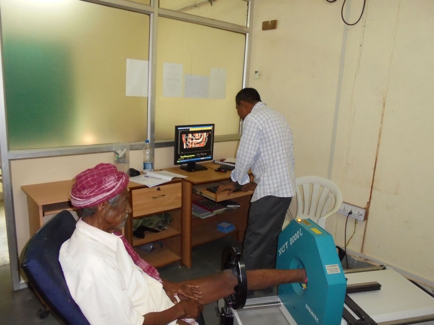 Technician operating diagnostic imaging equipment while an elderly patient undergoes lower limb scan in a clinical setting