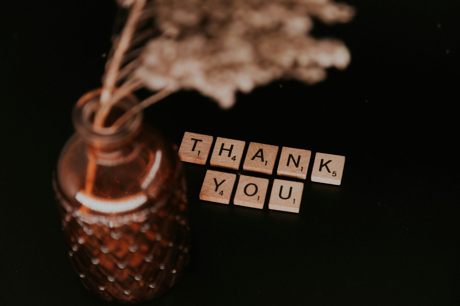 Decorative setup with wooden letter tiles spelling ‘Thank You’ beside a glass vase with dried flowers on a dark background