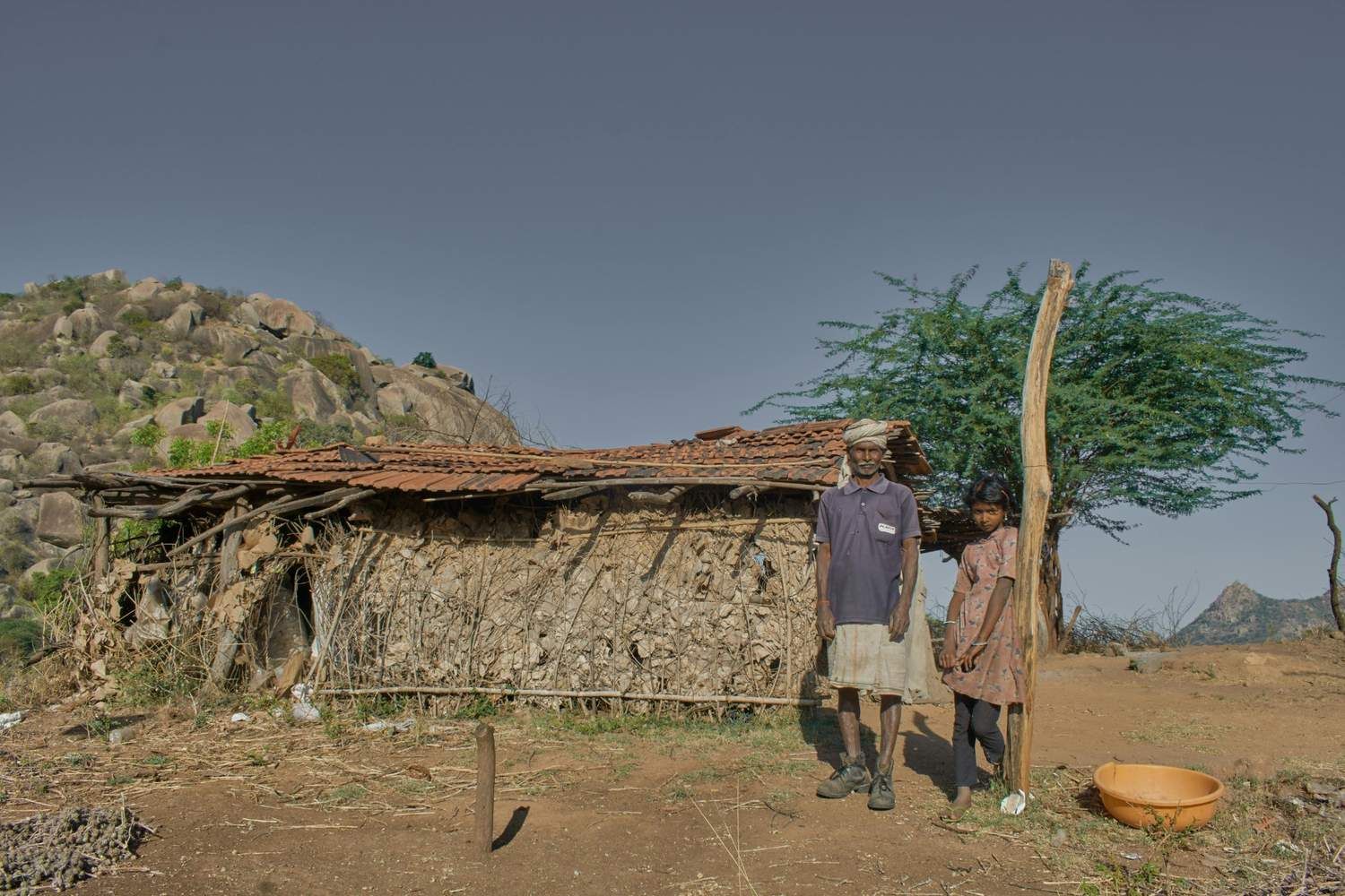An elderly man standing with his daughter outside their mud house.